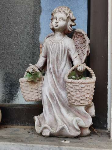 Une statue d'ange en pierre tenant deux paniers suspendus trône sur un rebord. L'ange porte une robe fluide, et de petites plantes vertes remplissent chaque panier.