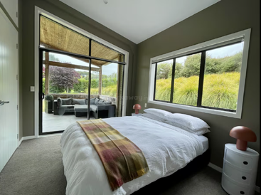 Dieses Schlafzimmer verfügt über große Fenster, ein gemütliches Bett mit weißer Bettwäsche, bunte Lampen und eine karierte Tagesdecke und öffnet sich zu einer Terrasse mit Sitzgelegenheiten im Freien und üppigem Grün.