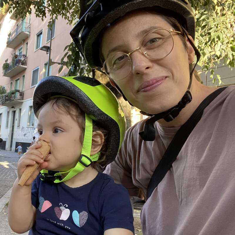 A woman and a child wearing helmets enjoy a sunny day outside. The child eats an ice cream cone, and they both look happy and relaxed.