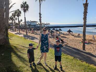 A woman and two young boys stand smiling on a grassy area near a sunny beach, with people relaxing under straw umbrellas and palm trees in the background.