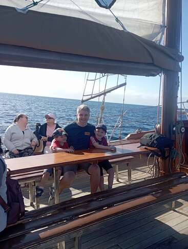 A family enjoys a sunny day sitting together on wooden benches aboard a sailboat, with calm blue ocean and clear sky in the background. Smiles and relaxation.
