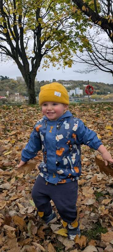 A smiling child in a yellow hat and blue jacket plays joyfully among autumn leaves in a park, surrounded by trees and distant buildings.