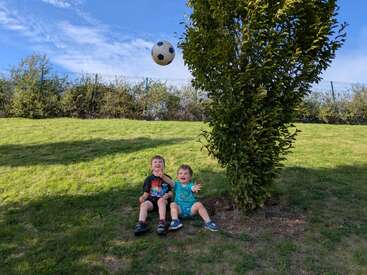 Two young children sit on green grass under a tree, laughing and playing. A soccer ball is mid-air above them. The sky is bright and blue.