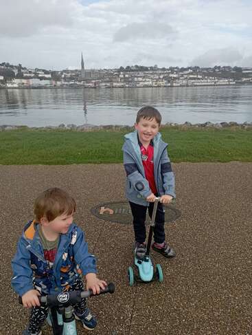 Two young boys on scooters smile by a waterfront path. Across the calm water, a picturesque town and church spire rise under a cloudy sky.