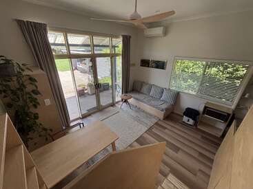 Esta imagem mostra uma sala de estar aconchegante e moderna, com janelas amplas, móveis de madeira clara, um sofá, uma mesinha, uma planta de interior e muita luz natural entrando.