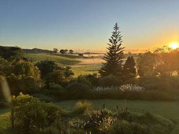 Um nascer do sol dourado banha uma paisagem verdejante com uma luz quente. Árvores altas, colinas onduladas e uma suave névoa matinal criam uma cena rural tranquila e serena.