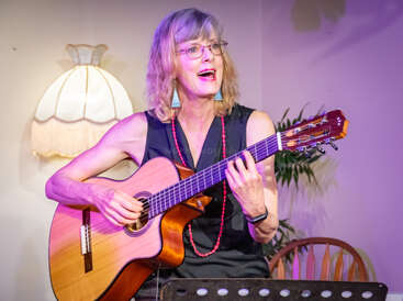 Una mujer rubia con gafas toca alegremente la guitarra acústica y canta. Lleva un vestido negro y un collar rojo, y actúa en una sala acogedora y con una iluminación cálida.