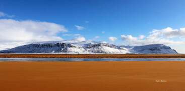 A vivid landscape features golden sandy foreground, a narrow river, and majestic snow-covered mountains beneath a clear blue sky with scattered fluffy clouds on horizon.
