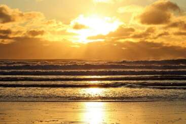 La imagen capta una hermosa puesta de sol sobre el océano, con la luz dorada del sol reflejándose en el agua, las olas ondulando y espectaculares nubes dispersas por el cielo.