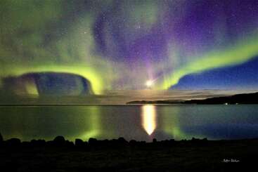 La imagen muestra impresionantes auroras boreales en tonos verdes y morados bailando sobre un lago en calma. Las estrellas brillan y la luna se refleja en el agua.