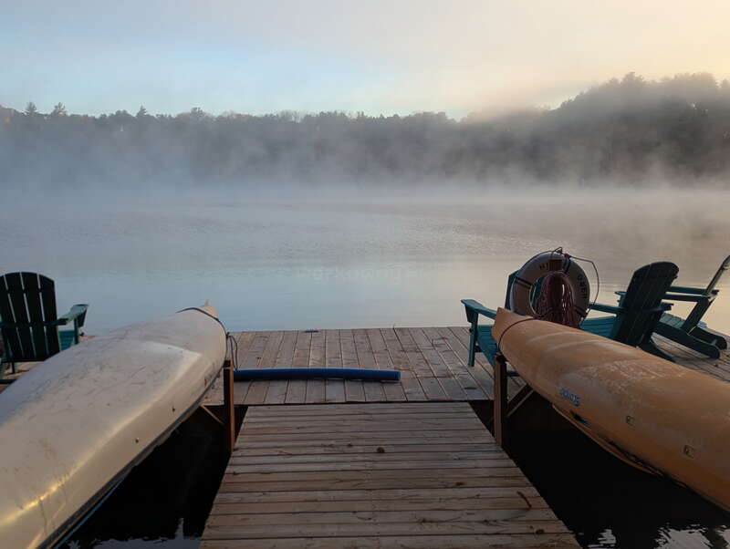 Un embarcadère paisible s'avance sur une eau calme et brumeuse. Deux kayaks sont amarrés de chaque côté, tandis que des chaises Adirondack et une bouée de sauvetage invitent à la détente.