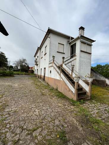 Uma casa branca de dois andares com escadas externas e persianas fechadas fica em uma entrada de pedra rachada, cercada por vegetação e sob um céu nublado.