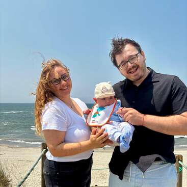 Una pareja sonriente sostiene a su bebé en la playa. El mar está de fondo y todos parecen felices, disfrutando juntos de un día soleado y ventoso.