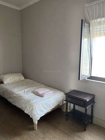 Um quarto simples com uma cama de solteiro, roupa de cama com estampa clara, toalha rosa dobrada, mesa de cabeceira de madeira, piso de madeira, paredes cinza lisas e uma janela aberta com cortinas transparentes.