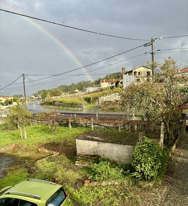 Um arco-íris vibrante se estende por um céu nublado sobre uma área rural com um carro verde, prédios antigos, estradas molhadas, vegetação exuberante, árvores e postes elétricos.