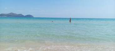 Une plage sereine et cristalline avec de douces vagues et une eau turquoise. Une personne se tient dans la mer peu profonde, des montagnes lointaines à l'horizon sous un ciel bleu clair.