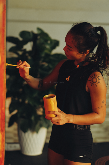 A woman with a sun tattoo paints a red surface while holding a yellow cup, focused and artistic, surrounded by a leafy indoor plant and warm lighting.