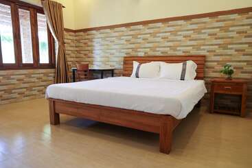 This image shows a spacious, clean bedroom with a wooden bed, white bedding, brick-patterned walls, a side table with a plant, desk, and large window.