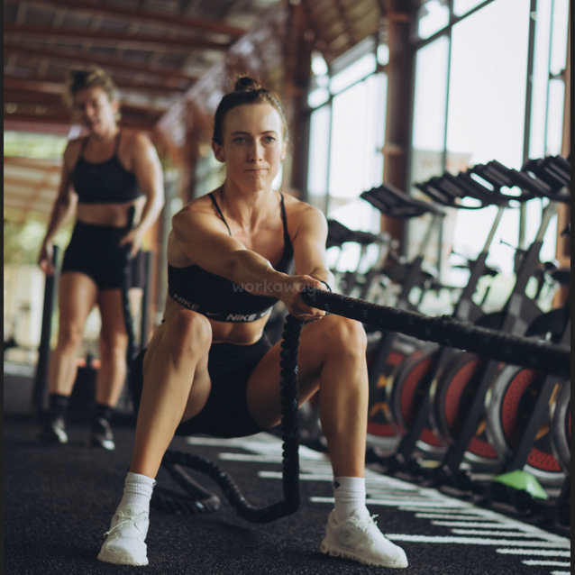 A woman in athletic wear exercises with battle ropes in a gym, focused and determined. Another woman in similar clothing is visible in the background, walking.