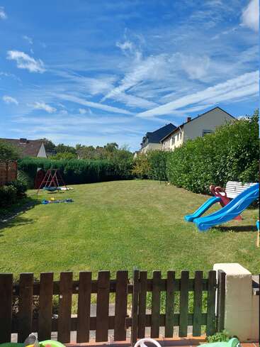 Une cour arrière ensoleillée avec une pelouse verte, des équipements de jeux pour enfants, notamment un toboggan et une balançoire, bordée de haies et de maisons, sous un ciel parsemé de nuages vaporeux.