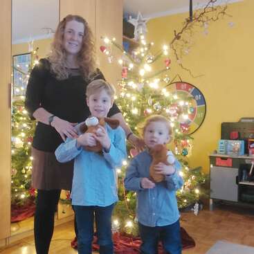Une femme et deux enfants se tiennent devant un sapin de Noël décoré et illuminé. Les enfants tiennent des peluches dans leurs bras, et la pièce est décorée de manière chaleureuse.