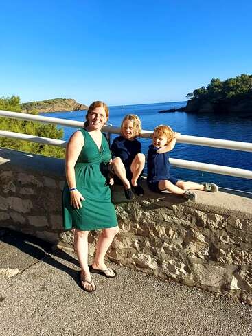 Una mujer con un vestido verde está de pie junto a un muro de piedra con dos niños pequeños, sonriendo y disfrutando de una pintoresca vista del mar bajo un cielo azul claro.