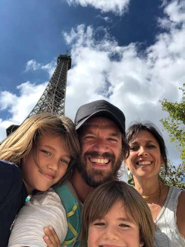 A joyful family poses together in front of the Eiffel Tower on a sunny day, smiling warmly with blue sky and fluffy clouds above them.