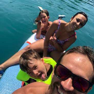 A happy family enjoys a sunny day on the water, smiling, posing on a paddleboard. Two kids give thumbs up while the father takes a selfie.