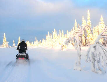 Eine Person fährt mit einem Schneemobil durch eine verschneite Landschaft, umgeben von schneebedeckten Bäumen, die von goldenem Sonnenlicht unter einem teilweise bewölkten Himmel wunderschön beleuchtet werden. Ein friedliches Winterabenteuer.