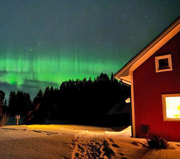 Eine gemütliche rote Hütte steht in einer verschneiten Landschaft, die von innen warm beleuchtet ist. Das Nordlicht leuchtet grün am Sternenhimmel und die Silhouetten der Wälder vervollständigen die ruhige Szene.