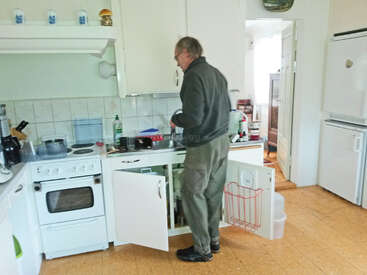 Ein Mann steht in einer Küche und wäscht Geschirr an der Spüle. Die Schranktüren sind geöffnet, und verschiedene Küchengeräte, Utensilien und Reinigungsmittel sind zu sehen.