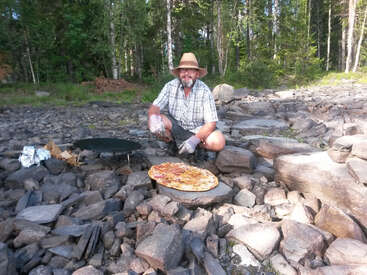 A man wearing a straw hat and gloves cooks a large pizza outdoors on a rocky surface, surrounded by trees, enjoying a rustic meal in nature.