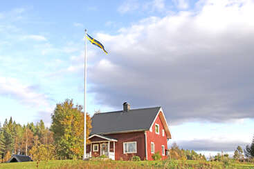 A red house with white trim stands amid greenery and trees. A Swedish flag flies on a tall pole. Clouds and blue sky fill the background.
