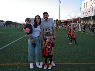 A family of four stands on a soccer field. The mother holds a young child, while the father stands beside their older child in a soccer uniform.