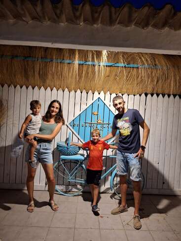 A family of four poses happily at night in front of a white fence with a blue bicycle and sign, under a straw-covered roof.