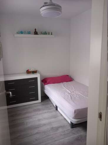 This is a small, minimalistic bedroom with a single bed, pink pillow, dresser, shelf with decorative items, white walls, and gray wooden flooring.