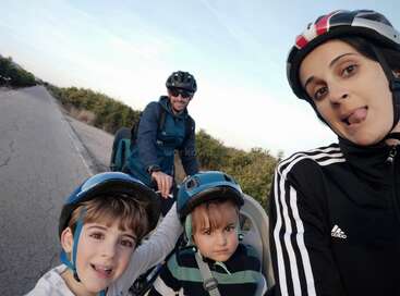 A family of four poses for a fun selfie during a bike ride. They’re all wearing helmets and smiling on a quiet road surrounded by greenery.