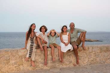 A family of five, barefoot and relaxed, sits on a stone wall by the sea at sunset, enjoying a peaceful, scenic moment together with gentle smiles.