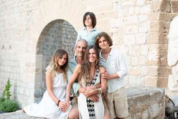 A happy family of five poses together outdoors by a stone wall and archway, smiling warmly. Two parents and three children appear close and joyful.