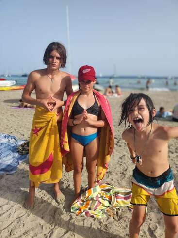 Three kids posing on a sunny beach. Two stand calmly with towels and serious faces, while the third is playful, sticking out his tongue enthusiastically. People relax behind.