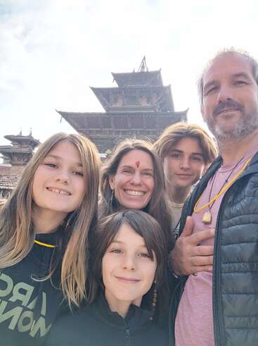 A smiling family takes a selfie in front of an ancient, multi-tiered temple. The background suggests they are traveling, exploring a historical or cultural site together.