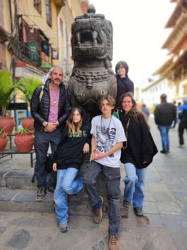 A group of five people poses around a traditional stone lion statue in a vibrant, historic street, surrounded by potted plants and old, colorful buildings.