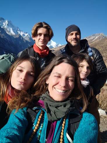 A family of five smiles outdoors, bundled warmly. Snow-capped mountains and clear blue sky form a stunning backdrop, suggesting a joyful hiking or trekking adventure.