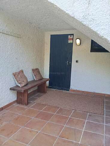 A cozy outdoor entryway features a wooden bench with two patterned pillows, a brown doormat, white textured wall, black door, coat hooks, and tiled floor.