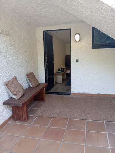 A cozy porch with terracotta tile floor, wooden bench with two patterned cushions, brown mat, white textured walls, open door, small window, and wall light.