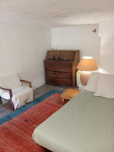 A cozy, simple room features a bed with white pillows, a wooden side table, armchair, large table lamp, chest of drawers, and a colorful rug.