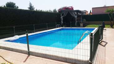 Una piscina rectangular está rodeada por una valla de seguridad. Hay una zona para sentarse a la sombra con cortinas al fondo, y árboles y vegetación alrededor del patio.