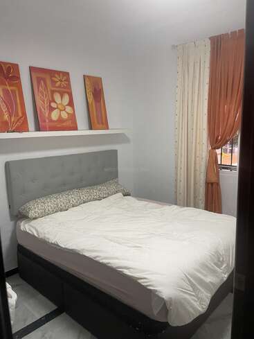 Este acogedor dormitorio presenta una cama bien hecha con un cabecero gris, obras de arte florales encima, paredes blancas y cortinas de color crema anaranjado junto a una ventana que proporciona una luz suave.