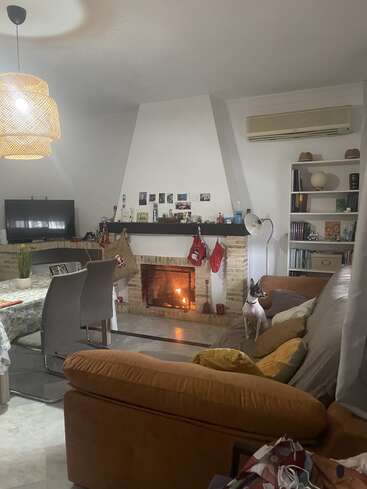 Un acogedor salón con chimenea encendida, medias, sofá marrón, perro en el sofá, mesa de comedor, televisor, estantería y una cálida iluminación decorativa, que crea un ambiente hogareño.