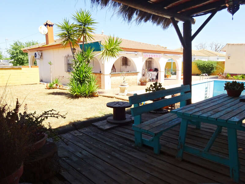 Un acogedor patio trasero con un sombreado patio de madera, un banco y una mesa de color turquesa, plantas en macetas, jardín de hierba seca, palmeras, piscina y un chalet blanco.
